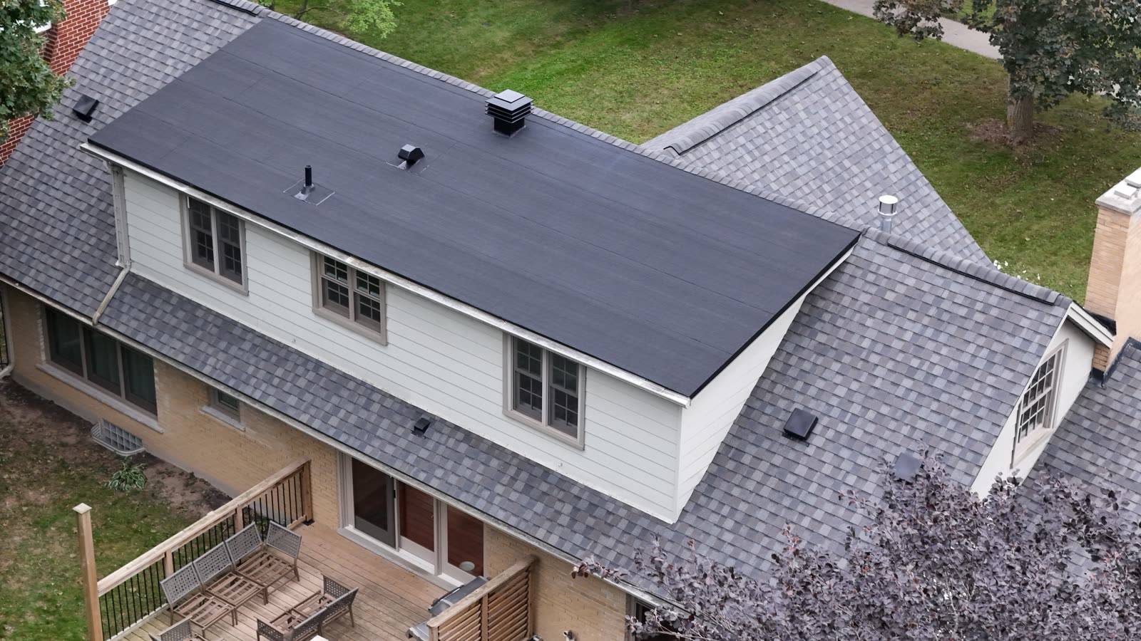Drone view of the back of a house: a flat membrane roof over a full-width dormer, surrounded by a finished Max Def Driftwood shingle roof